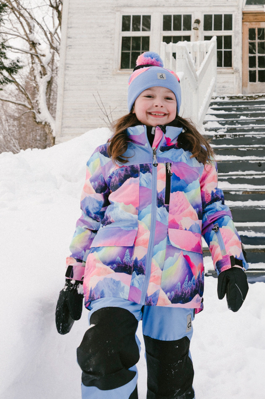 Child wearing the Deux par Deux PLAY two-piece snowsuit in Pink Mountain Print with Blue Pants during outdoor winter play