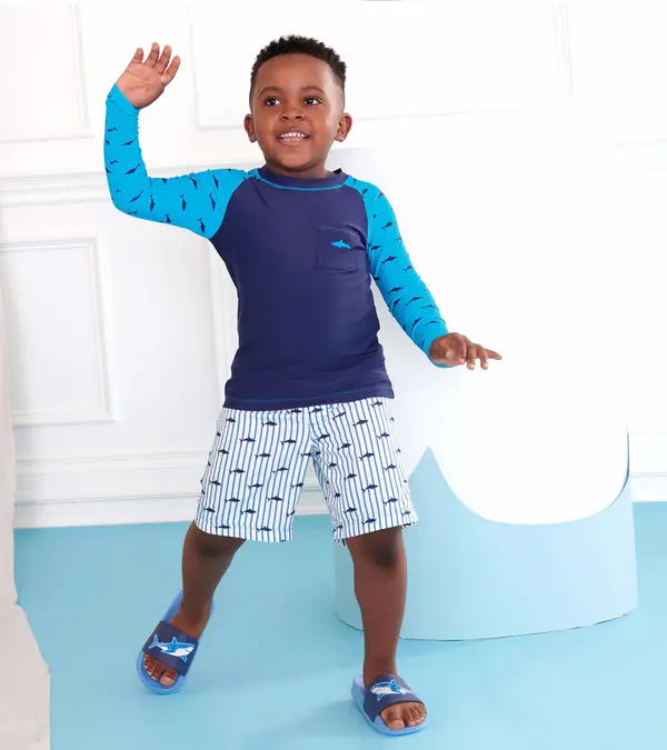 Little boy wearing blue shark board shorts with matching rashguard, standing by the pool.