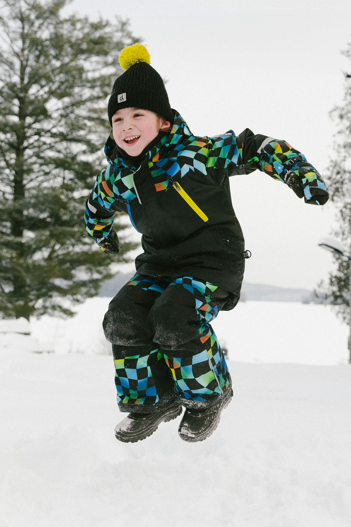 Boy wearing Deux par Deux checkerboard PLAY snowsuit jumping in snow