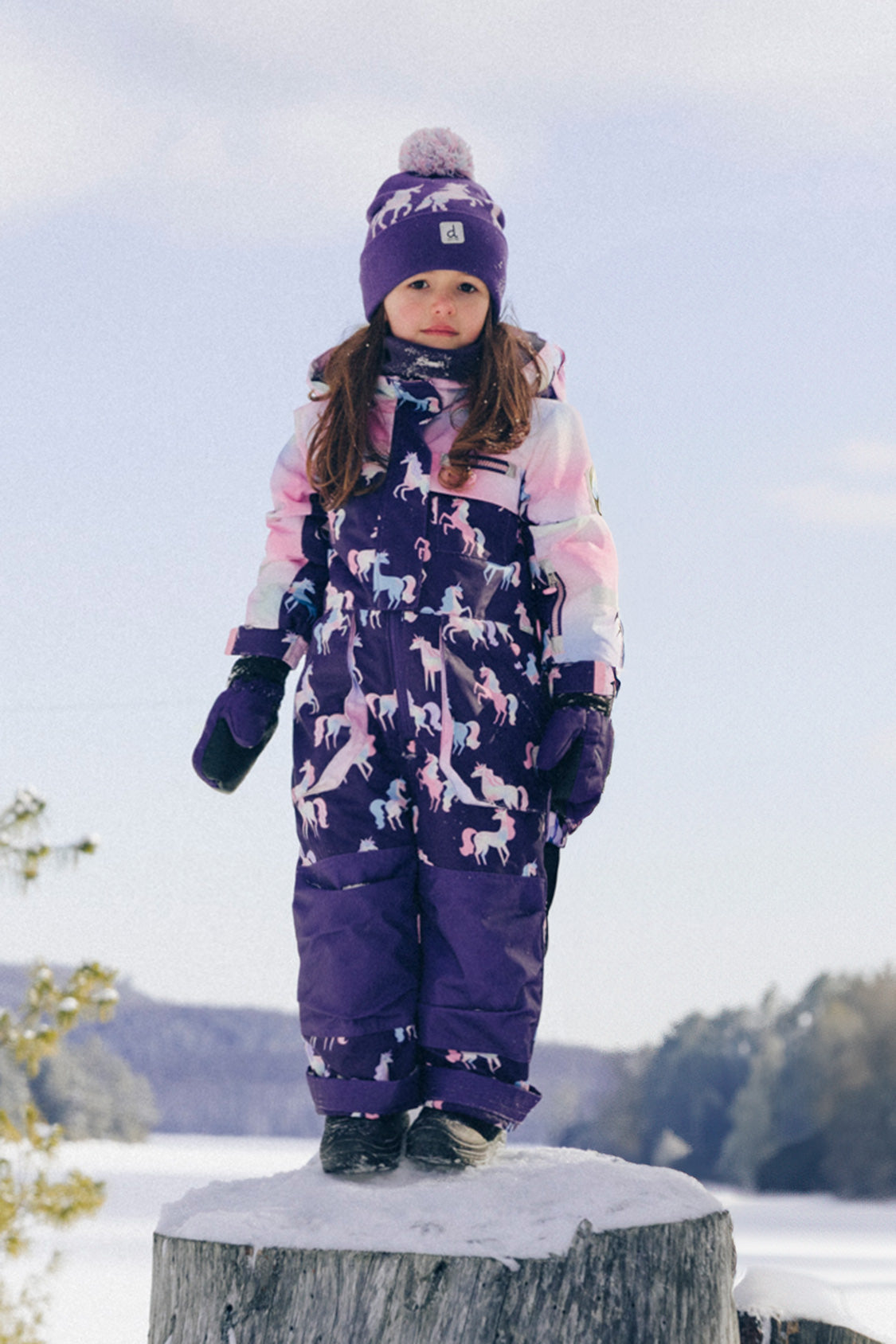 Child wearing the Deux par Deux kids PLAY one-piece snowsuit in Purple Unicorn Print during outdoor winter play