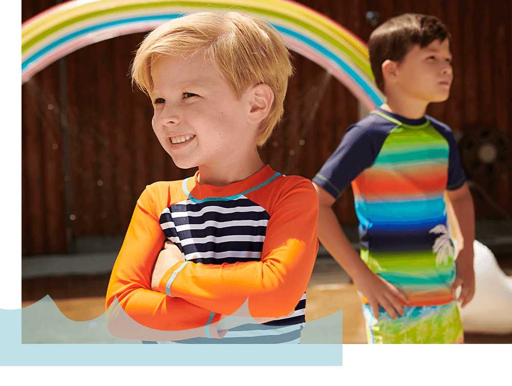 Boy wearing Hatley dino rash guard playing with rainbow sprinkler outside