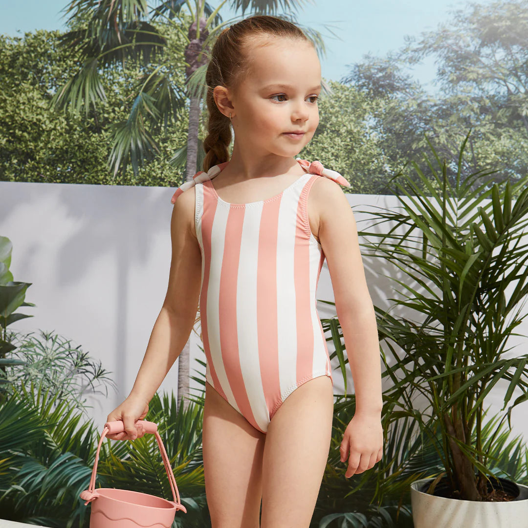 Girl wearing coral striped baby girl swimsuit standing by the pool with tropical plants.