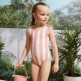 Girl wearing coral striped baby girl swimsuit standing by the pool with tropical plants.