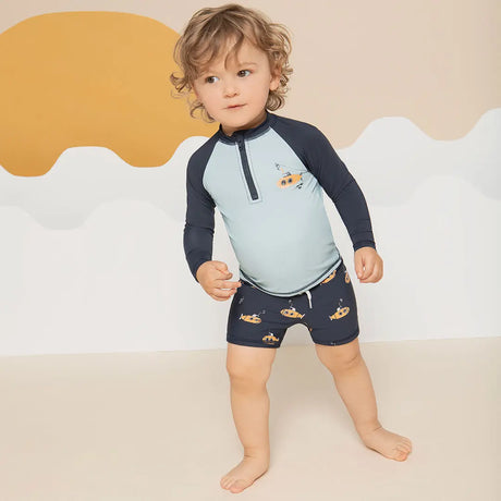 Petit Lem baby boy wearing navy raglan rash guard and striped submarine swim shorts on white background